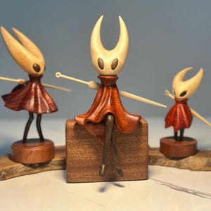 May include: Three wooden figurines of a character with a pale head and a red dress. The central figure sits on a wooden block, while the others stand on wooden bases. The figurines have outstretched arms and are set on a wooden base.