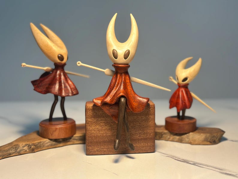 May include: Three wooden figurines of a character with a pale head and a red dress. The central figure sits on a wooden block, while the others stand on wooden bases. The figurines have outstretched arms and are set on a wooden base.