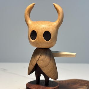 Peut inclure: Figurine en bois d'un personnage avec une t&ecirc;te cornue et une cape. La figurine a de grandes orbites sombres et un design simple et stylis&eacute;. La figurine est mont&eacute;e sur une petite base en bois ronde.