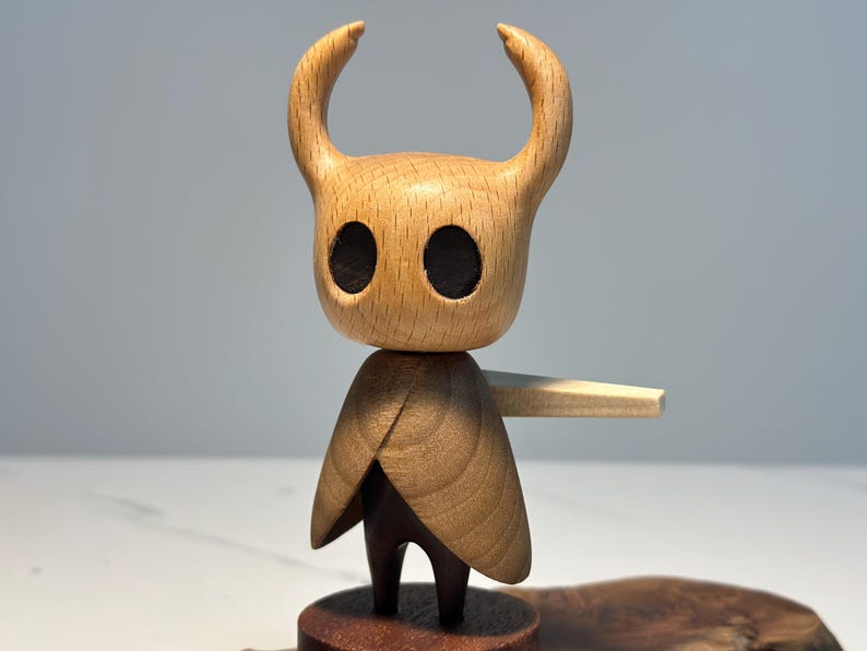 Peut inclure: Figurine en bois d'un personnage avec une t&ecirc;te cornue et une cape. La figurine a de grandes orbites sombres et un design simple et stylis&eacute;. La figurine est mont&eacute;e sur une petite base en bois ronde.