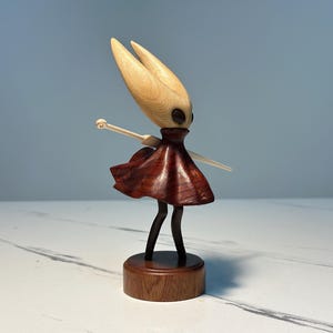 May include: A wooden figurine of a character with a light-coloured head and a dark red dress. The figure has black legs and is mounted on a round wooden base. The arms are outstretched, and the figure has a stylised appearance.