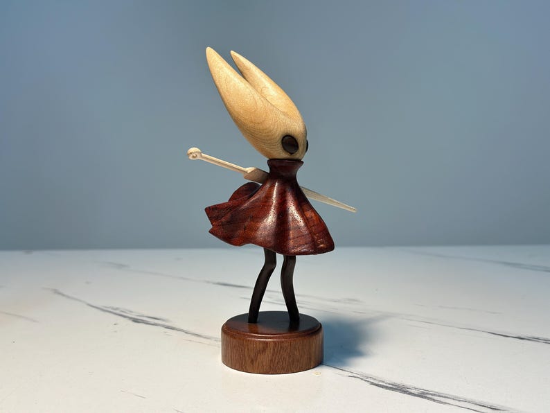 May include: A wooden figurine of a character with a light-coloured head and a dark red dress. The figure has black legs and is mounted on a round wooden base. The arms are outstretched, and the figure has a stylised appearance.