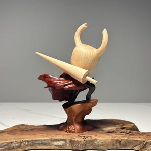 Peut inclure: Sculpture en bois d'un personnage avec une t&ecirc;te cornue et une grande arme pointue. La sculpture est faite de tons de bois clairs et fonc&eacute;s, avec un accent rouge. Elle est mont&eacute;e sur une base en bois.