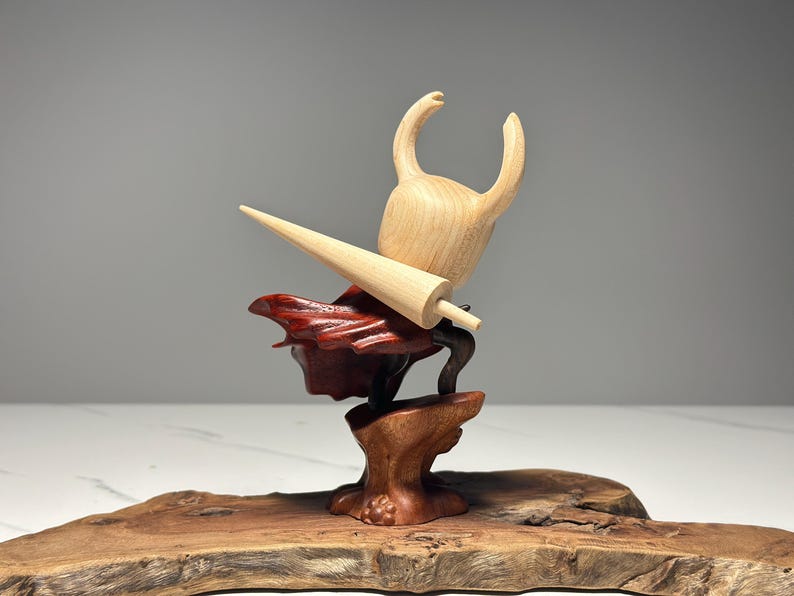 Peut inclure: Sculpture en bois d'un personnage avec une t&ecirc;te cornue et une grande arme pointue. La sculpture est faite de tons de bois clairs et fonc&eacute;s, avec un accent rouge. Elle est mont&eacute;e sur une base en bois.