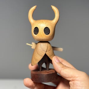 May include: Wooden figurine of a character with a horned head, black eyes, and a cape. The figure is made of light and dark brown wood and stands on a small, round base. The figurine is approximately 4 inches tall.