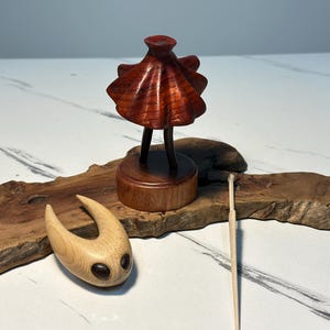 May include: A wooden sculpture of a figure with a flared top and two legs, on a round base. Another wooden sculpture with a curved shape and two dark eye-like features is also present. A thin wooden stick rests nearby.