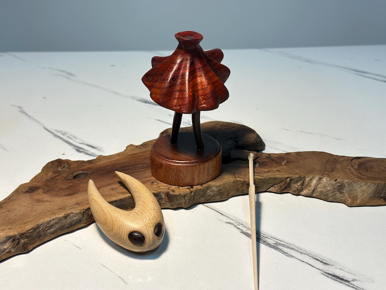 May include: A wooden sculpture of a figure with a flared top and two legs, on a round base. Another wooden sculpture with a curved shape and two dark eye-like features is also present. A thin wooden stick rests nearby.