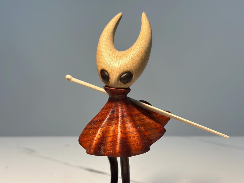 May include: A wooden figurine of a character with a light-coloured head and a reddish-brown skirt. The figurine has a light-coloured stick held across its body. The figure has black eyes and a dark brown neck.