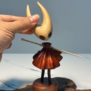 May include: A wooden figurine of a character with a light brown head and a dark brown body. The head has two large, dark eye-like features. The body has a skirt-like design. A hand is holding the head above the body.