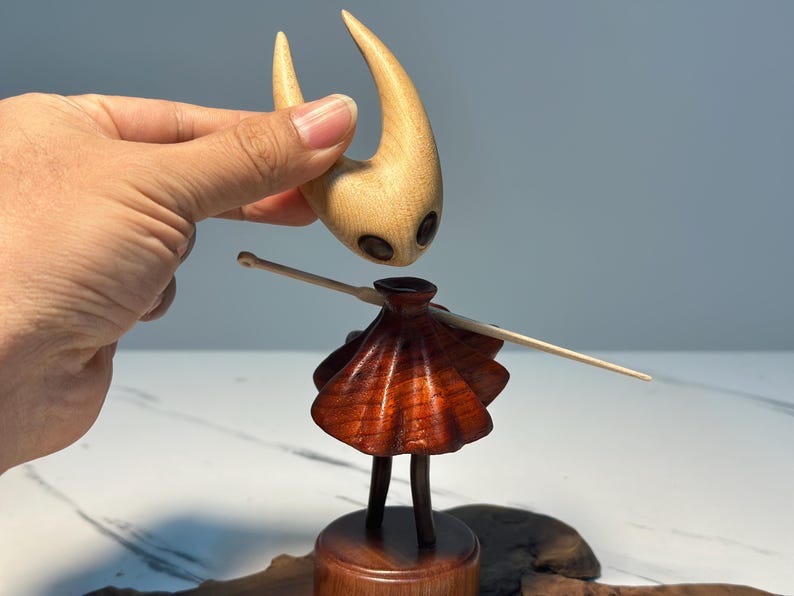 May include: A wooden figurine of a character with a light brown head and a dark brown body. The head has two large, dark eye-like features. The body has a skirt-like design. A hand is holding the head above the body.