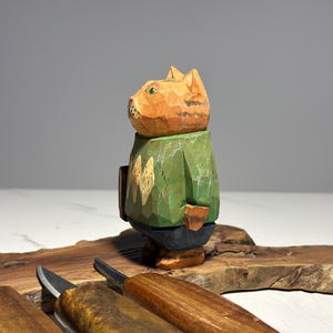 Może przedstawiać: Ręcznie rzeźbiona drewniana figurka kota z pomarańczowo-brązowym futrem, zielonymi oczami i zielonym swetrem. Kot stoi na drewnianej podstawie. Narzędzia do rzeźbienia na pierwszym planie.