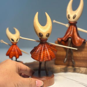 May include: Wooden figurines of a character with a light-coloured head and a red skirt. The figurines are in different sizes and are made of wood. The character has outstretched arms and is standing on a wooden base. The background is a light blue.