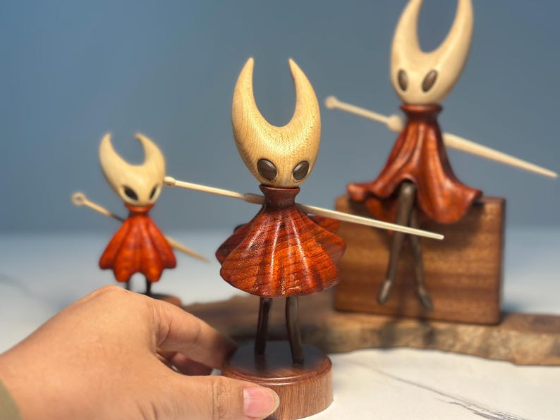 May include: Wooden figurines of a character with a light-coloured head and a red skirt. The figurines are in different sizes and are made of wood. The character has outstretched arms and is standing on a wooden base. The background is a light blue.