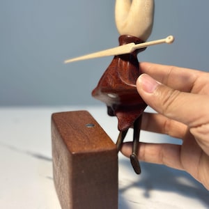 May include: A wooden figurine of a character with a light brown head and a dark red dress, holding a light brown sword. The figurine is positioned next to a rectangular wooden block. The figurine's legs are dark brown.