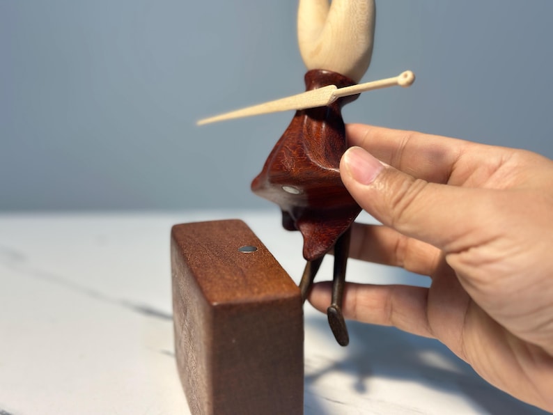 May include: A wooden figurine of a character with a light brown head and a dark red dress, holding a light brown sword. The figurine is positioned next to a rectangular wooden block. The figurine's legs are dark brown.