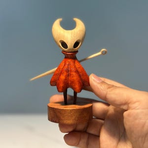 May include: A wooden figurine of a character with a light-coloured head and a red cloak. The figurine is mounted on a wooden base and holds a wooden needle. The character has two large, dark eye holes.