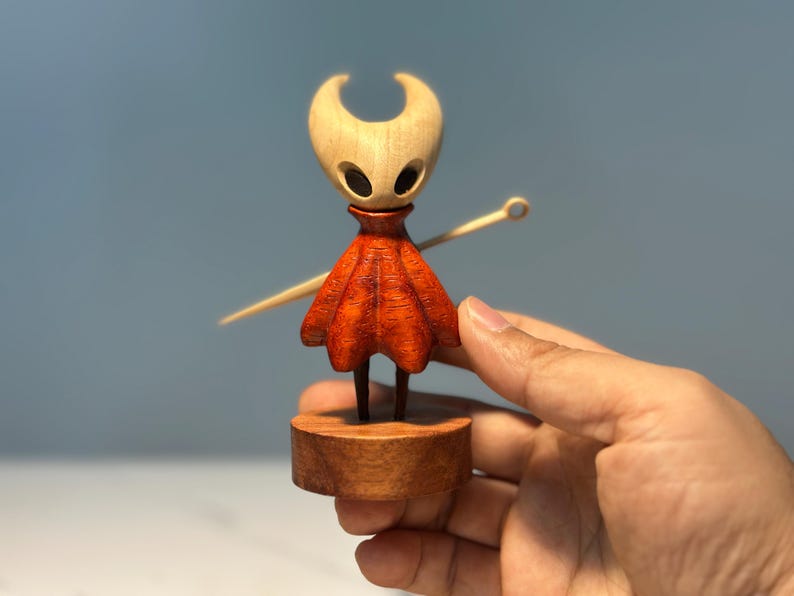 May include: A wooden figurine of a character with a light-coloured head and a red cloak. The figurine is mounted on a wooden base and holds a wooden needle. The character has two large, dark eye holes.