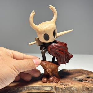 Peut inclure: Figurine en bois d'un personnage avec une t&ecirc;te de couleur claire, de grands yeux noirs et une corne pointue. La figurine tient une &eacute;p&eacute;e en bois et porte une tenue sombre et une cape rouge. La figurine est mont&eacute;e sur une base en bois.