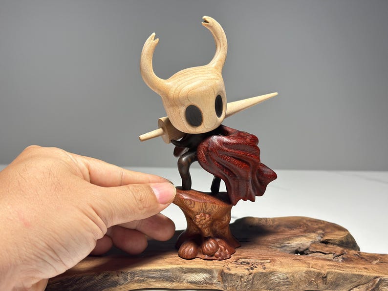 Peut inclure: Figurine en bois d'un personnage avec une t&ecirc;te de couleur claire, de grands yeux noirs et une corne pointue. La figurine tient une &eacute;p&eacute;e en bois et porte une tenue sombre et une cape rouge. La figurine est mont&eacute;e sur une base en bois.