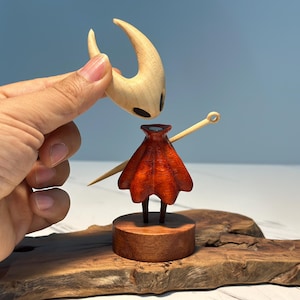 May include: A wooden figurine of a character with a light brown head and a red cape. The head is being held above the body. The figurine is on a wooden base and a wooden surface.