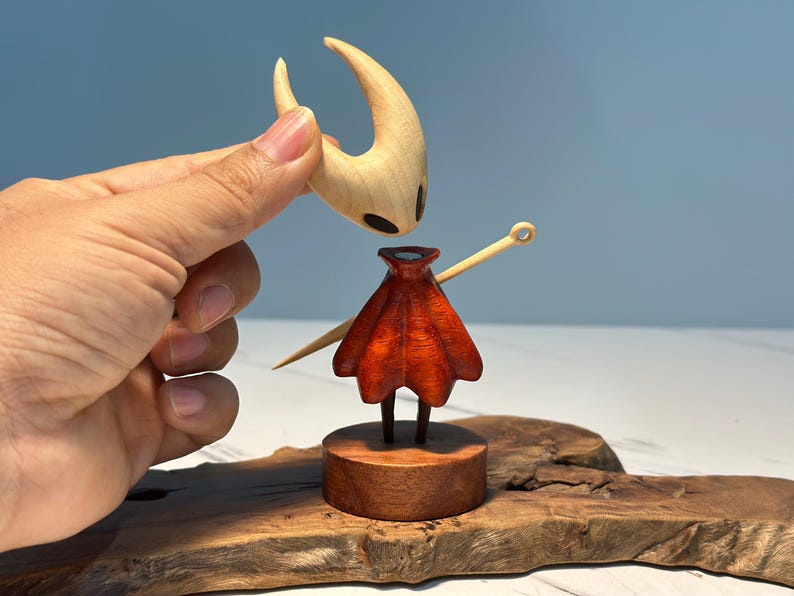May include: A wooden figurine of a character with a light brown head and a red cape. The head is being held above the body. The figurine is on a wooden base and a wooden surface.