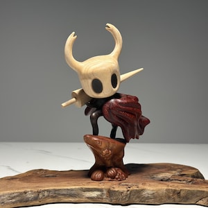 Peut inclure: Sculpture en bois d'un personnage avec une t&ecirc;te claire, de grands yeux noirs et des cornes incurv&eacute;es. Il porte une cape rouge fonc&eacute; et tient une arme en bois. La sculpture est mont&eacute;e sur une base en bois avec un bord naturel.
