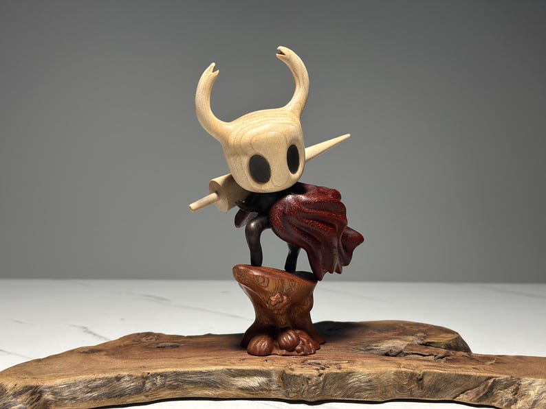 Peut inclure: Sculpture en bois d'un personnage avec une t&ecirc;te claire, de grands yeux noirs et des cornes incurv&eacute;es. Il porte une cape rouge fonc&eacute; et tient une arme en bois. La sculpture est mont&eacute;e sur une base en bois avec un bord naturel.