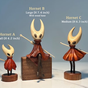 May include: Three wooden Hornet figurines of varying sizes. The smallest is 10.7 cm tall, the medium is 16 cm, and the largest is 18.8 cm with a wood base. Each figure has a light-colored head, a red skirt, and a wooden base.