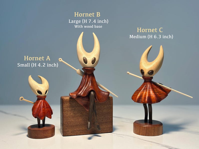 May include: Three wooden Hornet figurines of varying sizes. The smallest is 10.7 cm tall, the medium is 16 cm, and the largest is 18.8 cm with a wood base. Each figure has a light-colored head, a red skirt, and a wooden base.