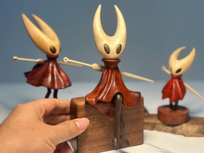 May include: Wooden figurines of a character with a pale head and a dark red dress. The figurines have outstretched arms and are set on wooden bases. The central figure is mounted on a wooden block. The background is a soft blue.