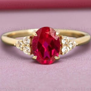 Puede incluir: Un anillo de oro con una gran piedra preciosa roja oscura de corte ovalado como pieza central. Pequeños grupos de piedras preciosas blancas flanquean la piedra principal. El anillo está sobre un fondo rosa suave.