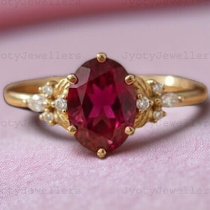 Op de afbeelding: Een gouden ring met een ovale, dieprode edelsteen als middelpunt. De ring is versierd met kleinere, heldere edelstenen en decoratieve gouden accenten. De ring wordt gepresenteerd tegen een roze achtergrond.