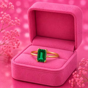 Peut inclure: Une bague en or ornée d'une pierre précieuse verte de taille émeraude rectangulaire montée sur quatre griffes. La bague est présentée dans un écrin à bijoux en velours rose vif ouvert, doté d'un motif en arc texturé sur la base avant.