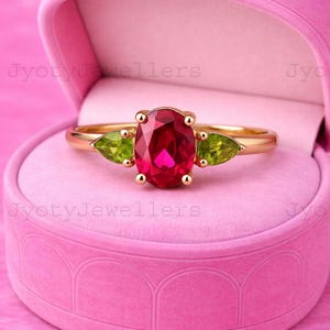 May include: A gold ring featuring a central oval-cut red gemstone flanked by two smaller pear-cut green gemstones. The ring is displayed inside a pink velvet jewelry box with the text JyotyJewellers visible on the box surface.