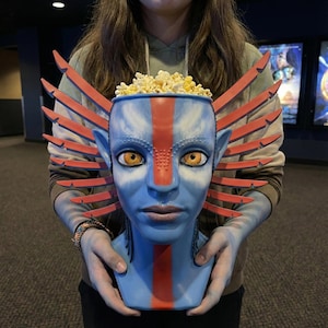 Pode incluir: Um balde de pipocas azul em forma de personagem Na'vi do filme Avatar, com olhos laranja e uma faixa vermelha no rosto. O balde está cheio de pipocas e tem detalhes em forma de asas vermelhas e laranja.