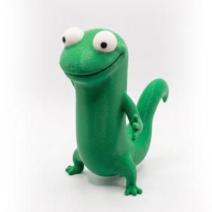 Könnte beinhalten: Eine hellgrüne, 3D-gedruckte Eidechsenfigur mit großen weißen Augen und einem lächelnden Gesichtsausdruck. Die Eidechse hat einen langen Schwanz und ist so positioniert, als würde sie gehen. Die Figur steht vor einem weißen Hintergrund.