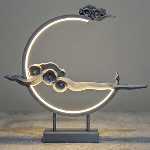May include: A gray decorative lamp featuring a crescent moon shape with a bright white light. The lamp has cloud-shaped accents and a small figure standing on a platform. The lamp is supported by a rectangular base.