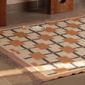 May include: A rectangular rug with a geometric pattern of brown squares and black lines on a beige background. The rug has a pink border and fringe. nursery rug, custom size runner, wool jute rug, navajo pattern rug, rustic modern rug, bedroom area rug, boho floor rug, hallway runner rug, living room area rug, red geometric rug, red handwoven rug, red southwestern rug, red stairs runner, vintage red rug