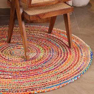 Könnte beinhalten: Ein runder, mehrfarbiger geflochtener Jute-Teppich mit einer lebendigen Farbpalette aus Rot, Orange, Gelb, Grün und Blau. Der Teppich liegt teilweise unter einem Holzstuhl mit gewebter Sitzfläche. Der Text "Rug creation IN" ist sichtbar.