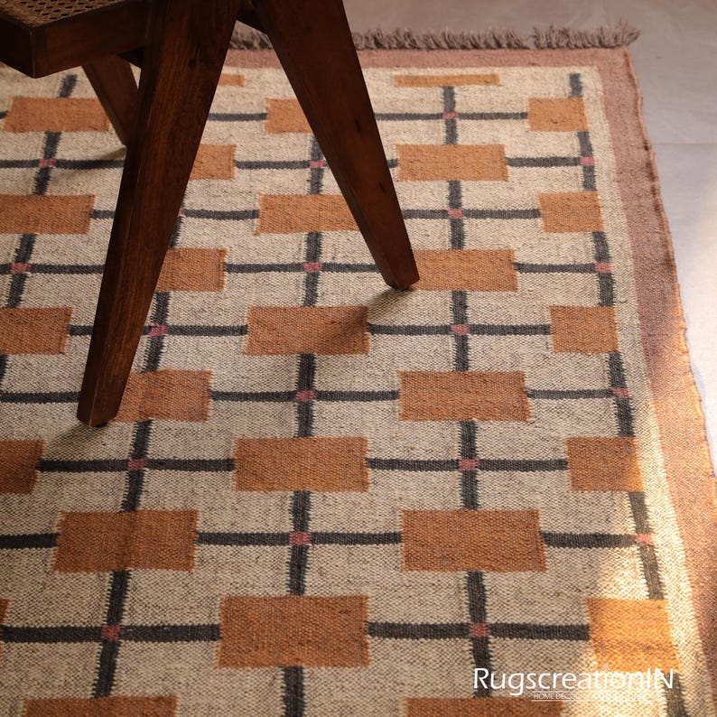 Op de afbeelding: Een beige tapijt met een geometrisch patroon van bruine rechthoeken en zwarte lijnen. Het tapijt heeft een bruine rand en een houten stoelpoten zijn zichtbaar. Het tapijt is gemaakt van geweven materiaal. Afmetingen in centimeters.