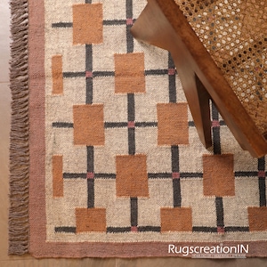 Op de afbeelding: Beige tapijt met een geometrisch patroon van bruine vierkanten en zwarte lijnen, met roze accenten. Het tapijt heeft een franjerand. Een houten stoel met een geweven zitting is gedeeltelijk zichtbaar. De tekst "RugscreatioIN" is zichtbaar.