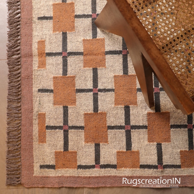 Op de afbeelding: Beige tapijt met een geometrisch patroon van bruine vierkanten en zwarte lijnen, met roze accenten. Het tapijt heeft een franjerand. Een houten stoel met een geweven zitting is gedeeltelijk zichtbaar. De tekst "RugscreatioIN" is zichtbaar.