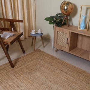 Puede incluir: Una alfombra rectangular de yute con un diseño de borde geométrico. La alfombra es de color marrón claro y está colocada sobre un suelo embaldosado. La habitación cuenta con muebles de madera y objetos decorativos.