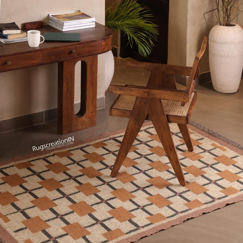 Op de afbeelding: Een patroonkleed met een beige achtergrond en bruine en zwarte vierkanten. Een houten stoel met een geweven zitting staat op het kleed. Een houten bureau en een witte mok staan op de achtergrond. Het kleed heeft een gefranjerde rand.