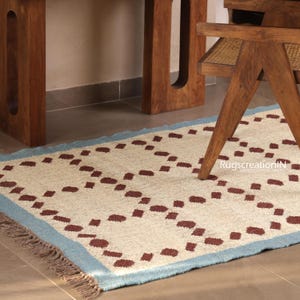 Puede incluir: Una alfombra rectangular con un centro color crema y un borde azul claro. La alfombra presenta un patr&oacute;n repetitivo de formas punteadas de color marr&oacute;n oscuro. La alfombra est&aacute; colocada sobre un suelo embaldosado, con una silla y una mesa de madera al fondo.