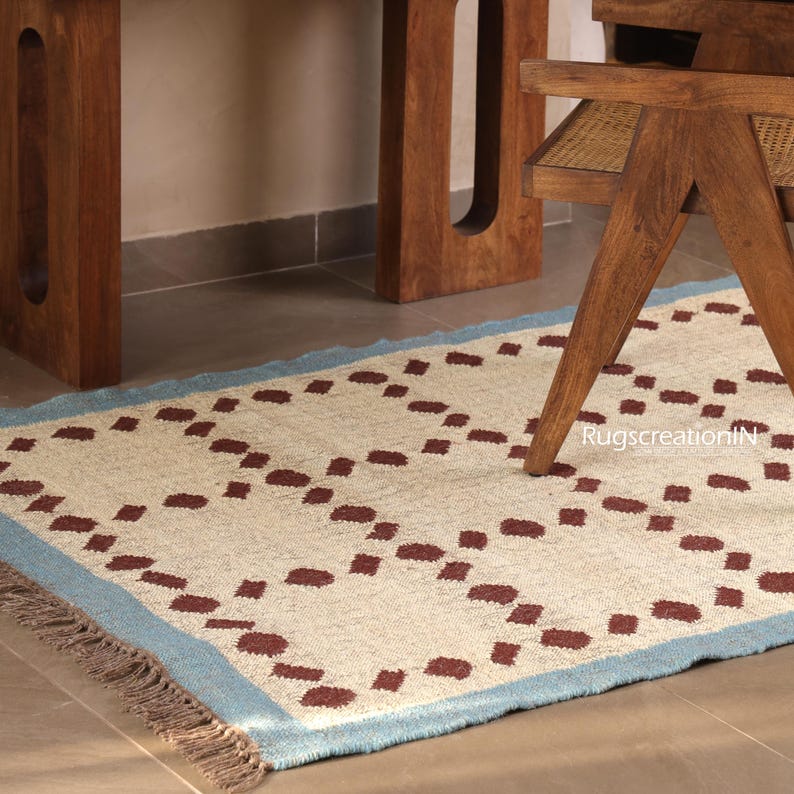 Puede incluir: Una alfombra rectangular con un centro color crema y un borde azul claro. La alfombra presenta un patr&oacute;n repetitivo de formas punteadas de color marr&oacute;n oscuro. La alfombra est&aacute; colocada sobre un suelo embaldosado, con una silla y una mesa de madera al fondo.