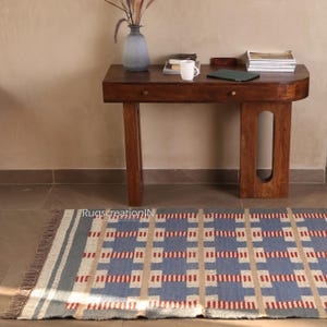 May include: A rectangular blue, beige, and red patterned rug with a fringed edge is placed on the floor. A dark wooden desk with two drawers and a vase with dried plants sits in the background. The desk has a curved front and a light-colored wall.