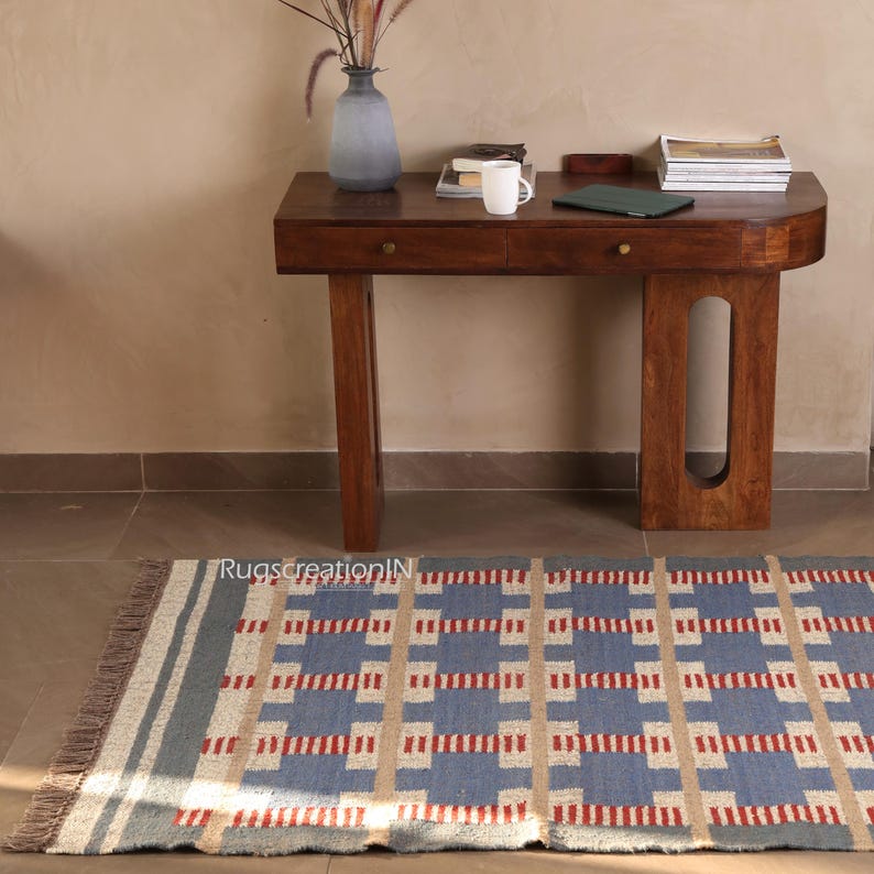 May include: A rectangular blue, beige, and red patterned rug with a fringed edge is placed on the floor. A dark wooden desk with two drawers and a vase with dried plants sits in the background. The desk has a curved front and a light-colored wall.