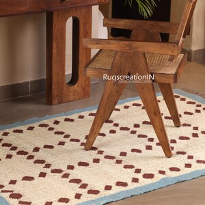Puede incluir: Una alfombra color crema con un patr&oacute;n repetitivo de cuadrados granates y un borde azul claro. Una silla de madera con asiento tejido y un escritorio de madera est&aacute;n en el fondo. La alfombra es rectangular y parece estar hecha de fibras naturales.