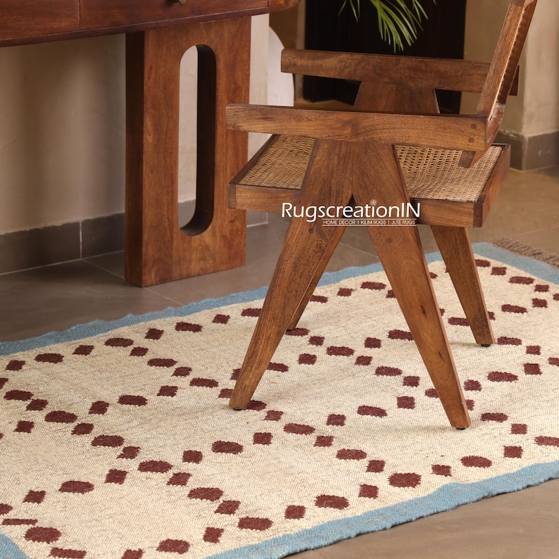 Puede incluir: Una alfombra color crema con un patr&oacute;n repetitivo de cuadrados granates y un borde azul claro. Una silla de madera con asiento tejido y un escritorio de madera est&aacute;n en el fondo. La alfombra es rectangular y parece estar hecha de fibras naturales.